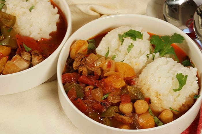 Two white bowls filled with stew. Each bowl has two dollops of rice and the stew is served alongside. The meal is garnished with chopped cilantro.