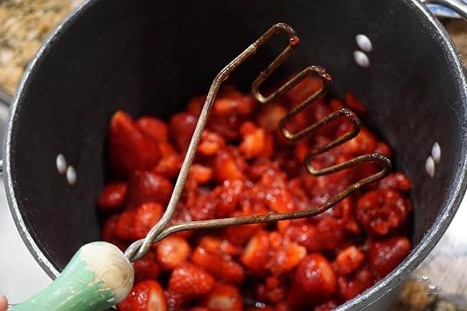A heavy black pot filled with berries getting mashed to make a strawberry preserves recipe