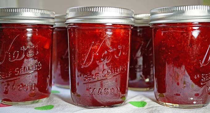 Clear jars of jam filled with a strawberry preserves recipe are lined up on a white towel and the jars are sealed.
