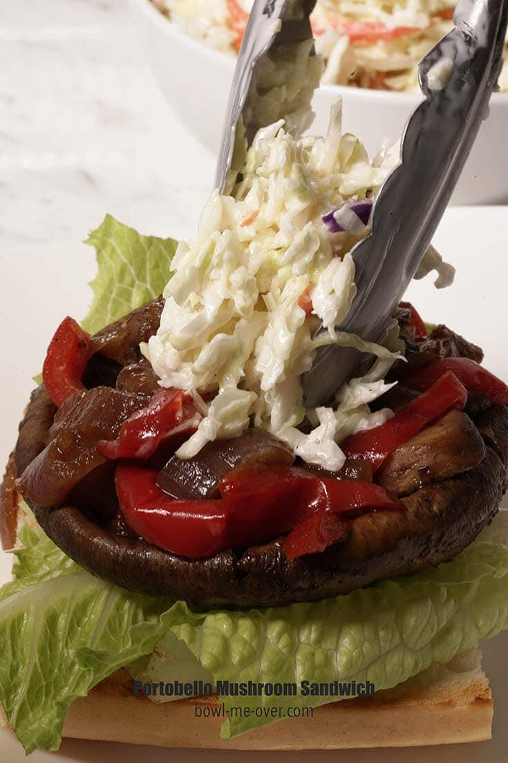 A burger bun topped with lettuce, portobello mushroom, onions and peppers. There's a spoonful of coleslaw being placed on the top.