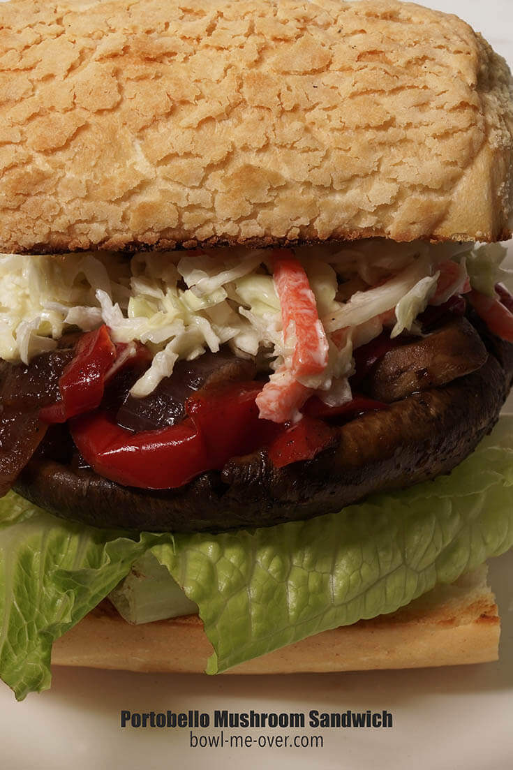 Close-up picture of a sandwich. The bottom bun is topped with lettuce, portobello mushroom, red peppers and onions. The sandwich is topped with crunchy coleslaw and finished off with a big bun on top.