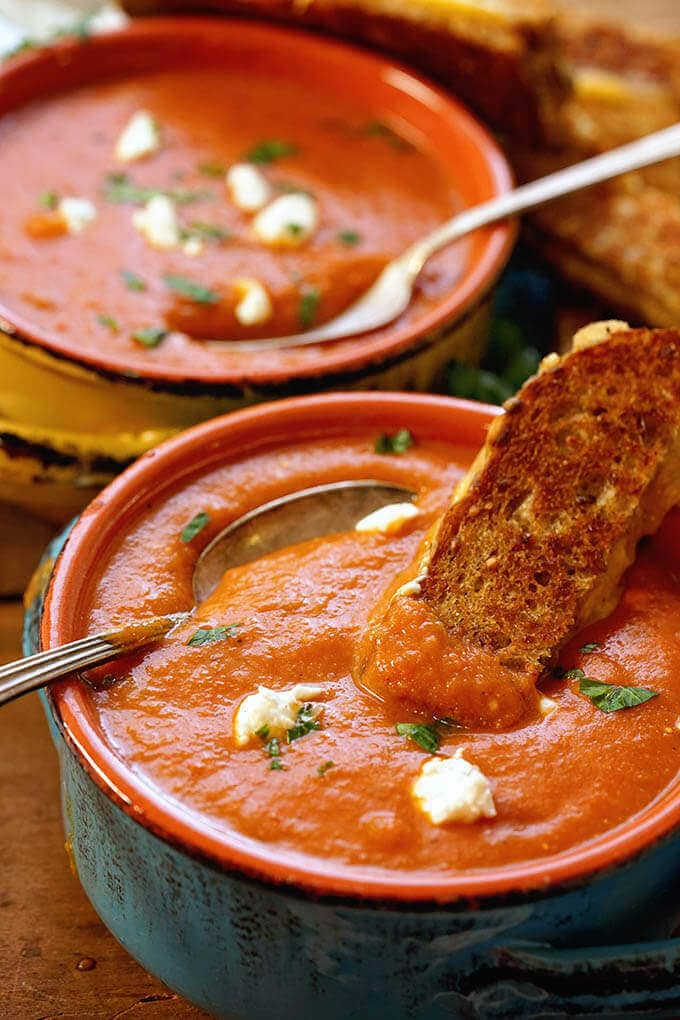 Two bowls of tomato bisque soup topped with goat cheese and served with grilled cheese sandwich.