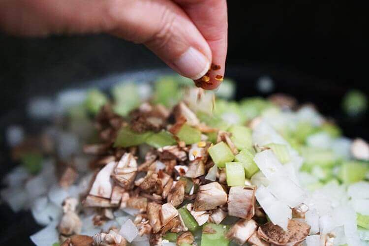Celery, mushrooms and onions in a cast iron skillet. A pinch of red pepper flakes is being added to the vegetables.