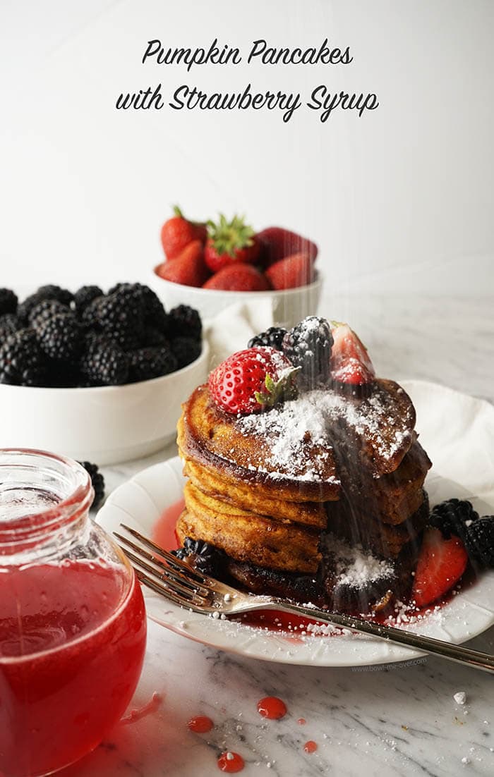 A short stack of golden brown pumpkin pancakes topped with strawberries and blackberries. It's been drizzled with strawberry syrup. There is extra syrup for passing and bowls of blackberries and strawberries in the background.