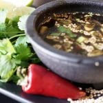 A black bowl filled with Citrus Ponzu Dipping Sauce. The bowl is surrounded by egg rolls, red chili pepper, cilantro and sesame seeds.