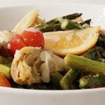 A white bowl filled with wedges of lemon, sliced tomatoes, marinated artichoke hearts and roasted asparagus.