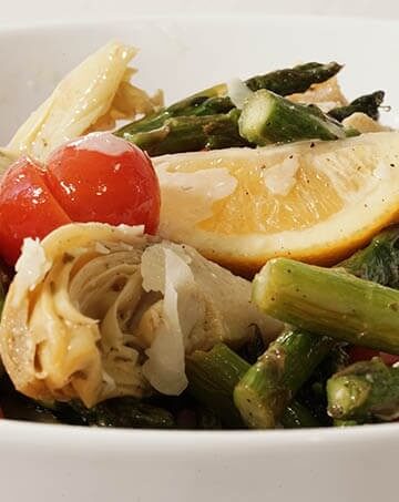 A white bowl filled with wedges of lemon, sliced tomatoes, marinated artichoke hearts and roasted asparagus.