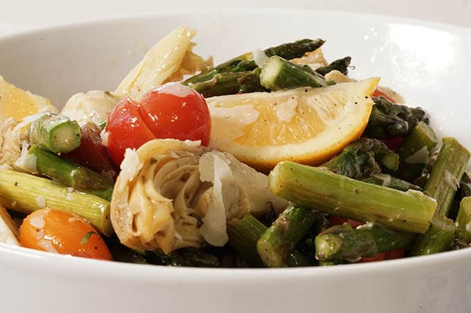 A white bowl filled with wedges of lemon, sliced tomatoes, marinated artichoke hearts and roasted asparagus.