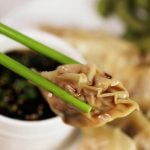A close up picture of green chopsticks holding a savory potsticker. There's a bowl of dipping sauce in the background and also a plate of Pot stickers to share.