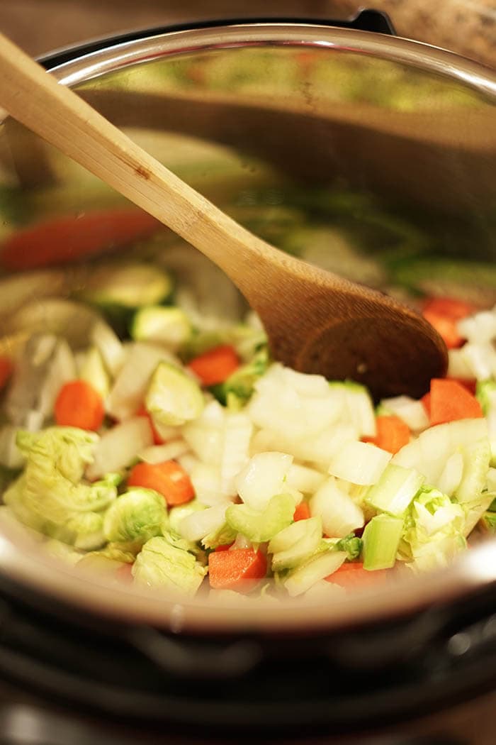 A picture of the inside of an Instant Pot. There are carrots, onions, and brussels sprouts sautéing and a wooden spoon stirring the vegetables.
