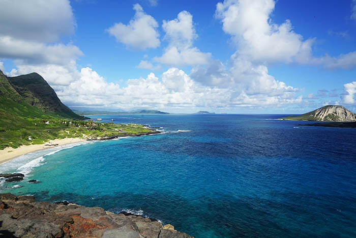 A breathtaking view from the lookout at Makapuu. High above the ocean the viewing showcases the green mountains in stark contract to the bright blue ocean. The beautiful blue sky is dotted with white puffy clouds.