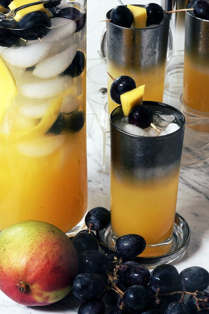 Glasses of mango punch topped with grapes and chunks of mango. The glasses are surrounded by bunches of grapes and fresh mango.