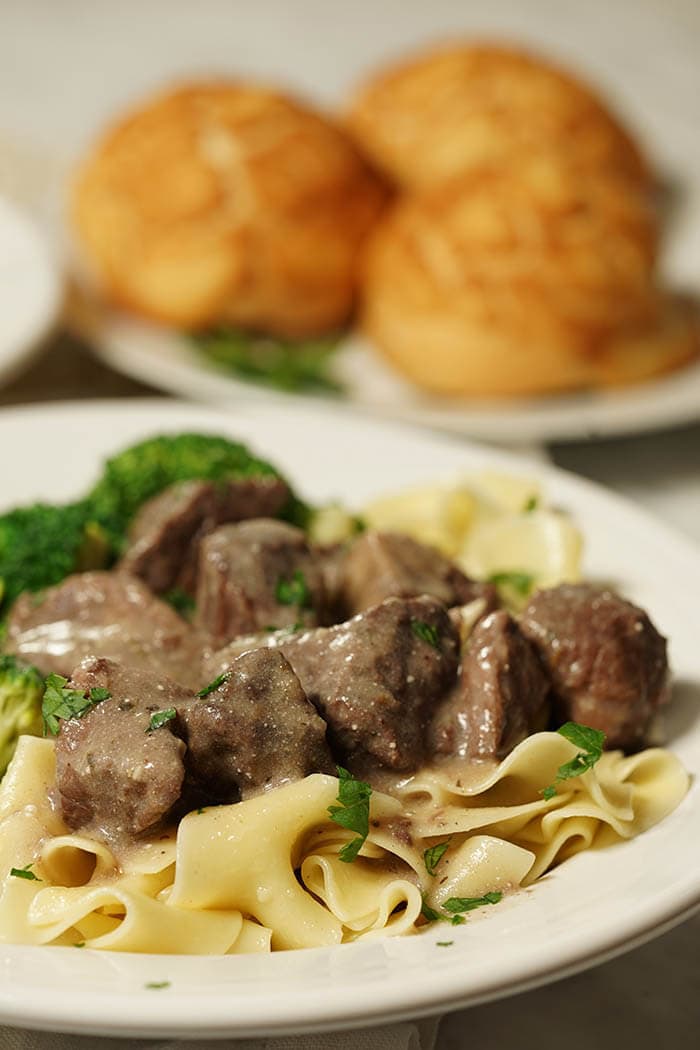 A white bowl full of noodles topped with tender beef tips. Served with a side of broccoli and fresh made rolls. Beef Tips with Gravy Recipe.