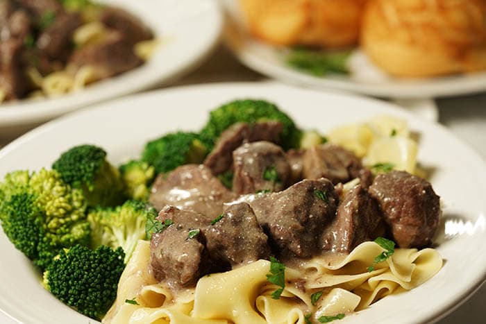 A white bowl full of egg noodles. The egg noodles are topped with a couple scoops of tender beef tips smothered in brown gravy.