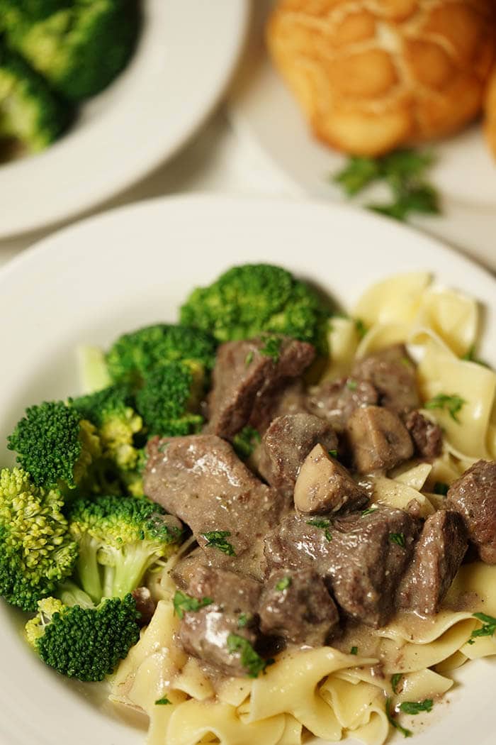A shallow white bowl heaped with egg noodles. The noodles are smothered in Beef Tips with Gravy. This meal is served with steamed broccoli and a roll.