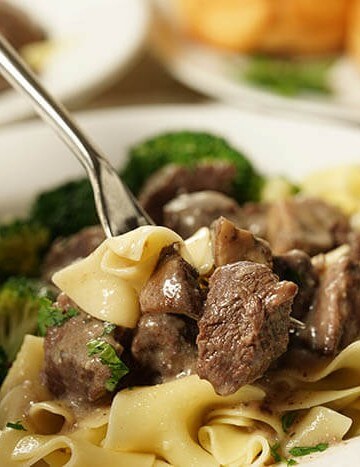 A white bowl filled with Beef Tips with Gravy served overtop noodles. A fork is digging in, ready to take a hearty bite! This meal is served with a roll and side of steamed broccoli.