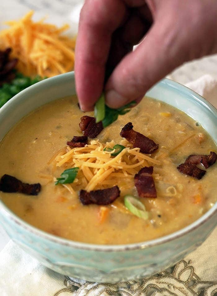 A green bowl full of corn and potato chowder topped with bacon and cheese. A hand is sprinkling thinly sliced green onions onto the top of the chowder.