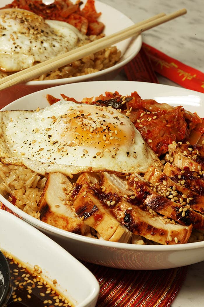A bowl with brown rice, topped with kimchi, a fried egg and grilled chicken with a Korean BBQ Sauce Recipe. The whole bowl is sprinkled with sesame seeds.