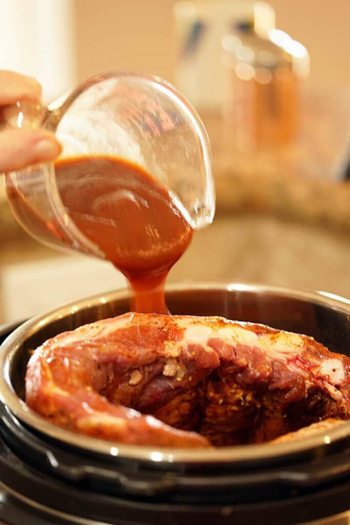 A pressure cooker with pork ribs rolled up and place in the pot. Barbecue sauce is being drizzled over the side.