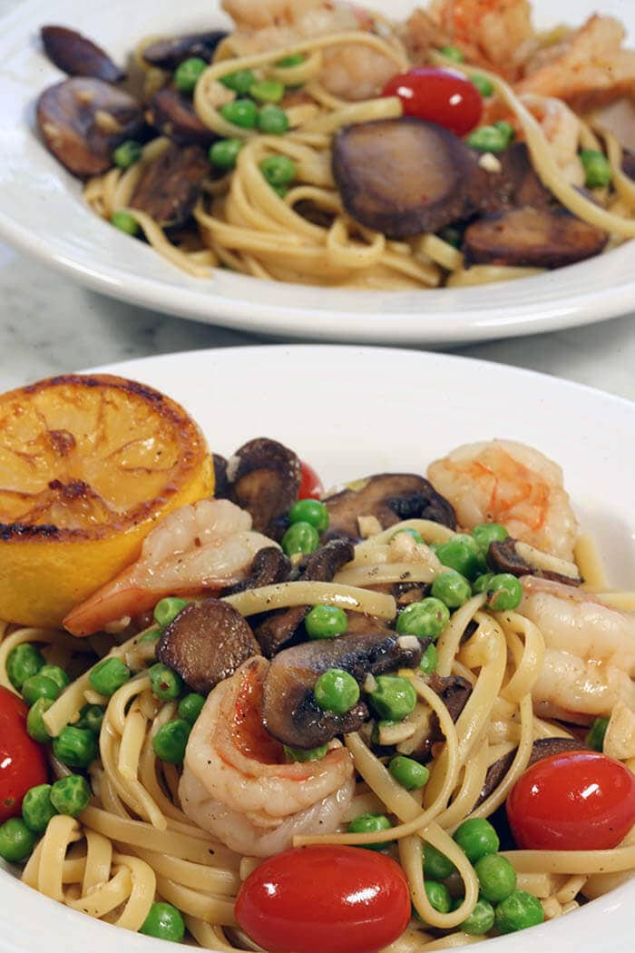 Two white bowls filled with Shrimp Scampi Linguine mixed with cherry tomatoes, sautéed mushrooms and bright green peas.