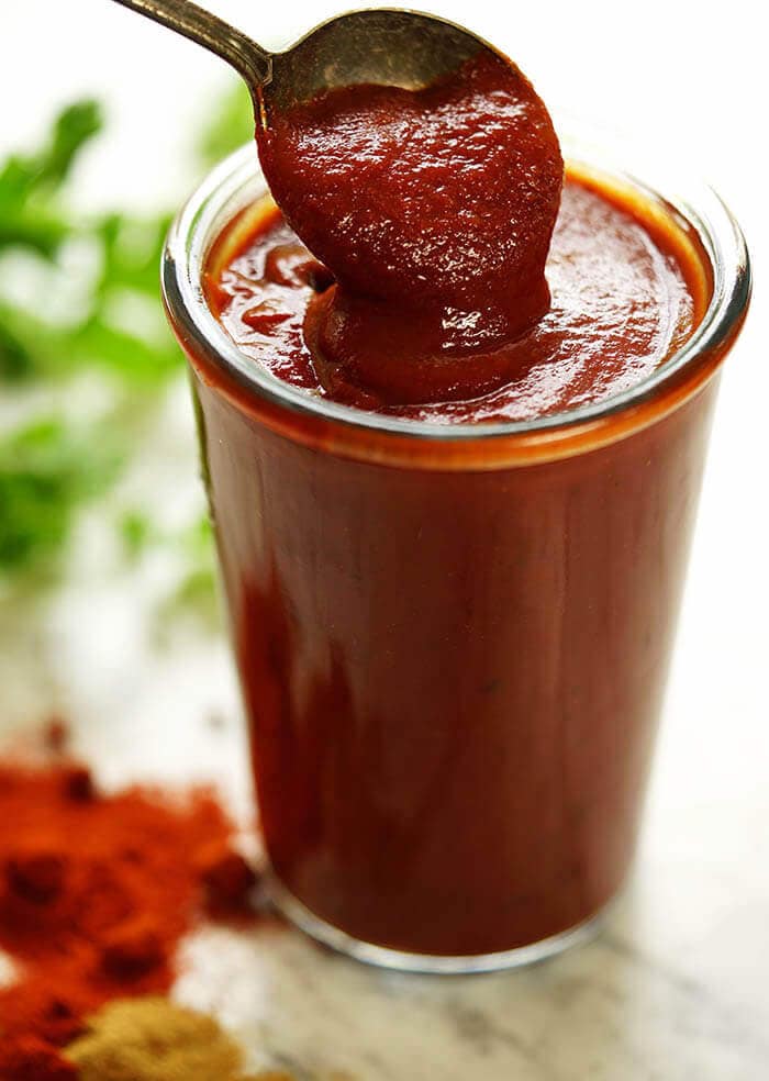 A glass jar filled with the best enchilada sauce surrounded by scattered spices and herbs