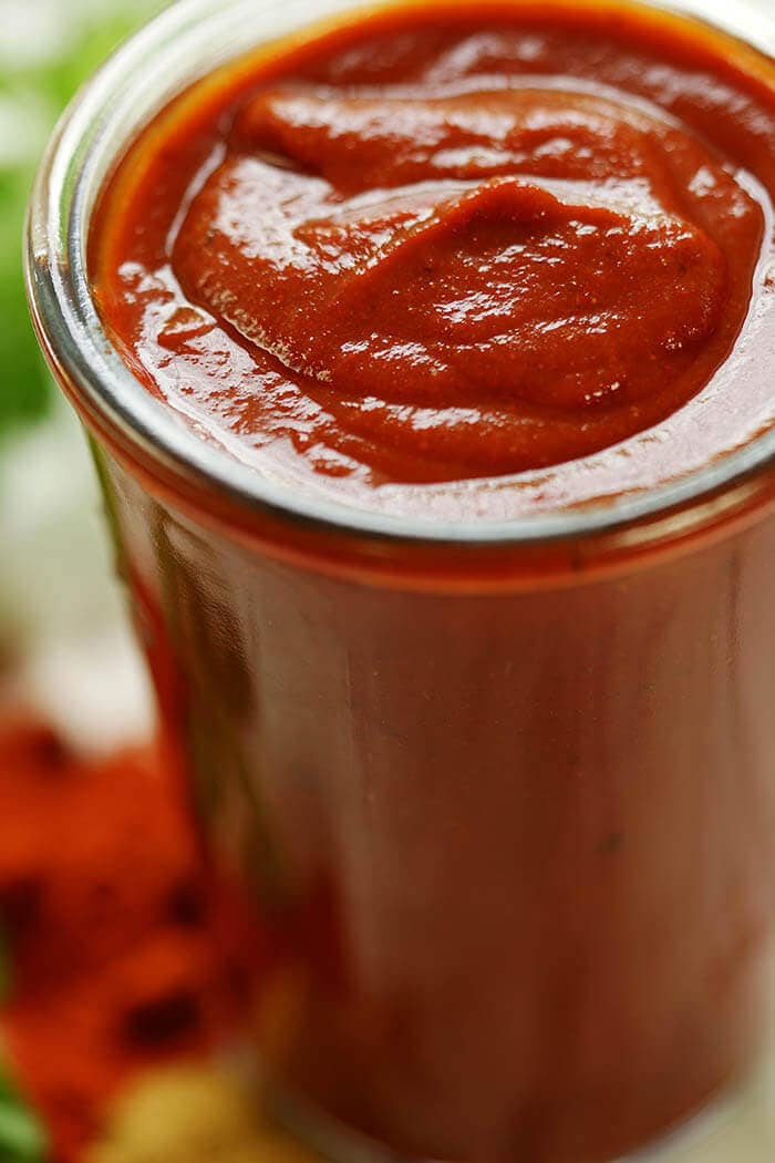 A jar of red chili enchilada sauce made with red chili powder and cumin. There's some cilantro in the background for garnish. 