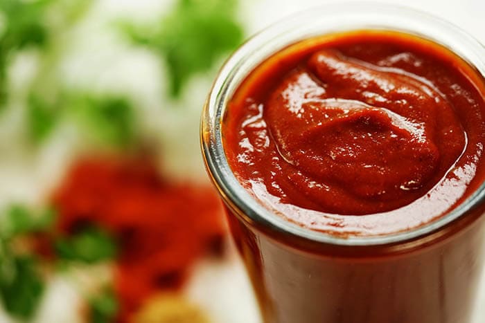 Red enchilada sauce in a glass jar surrounded by herbs and spices.