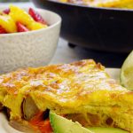 A white plate with a wedge of frittata served with a side of avocado and fresh fruit. This is a recipe for an easy frittata