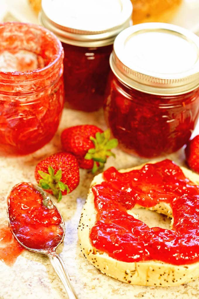 Jars of homemade strawberry jam surrounding a bagel spread with sweet jam!