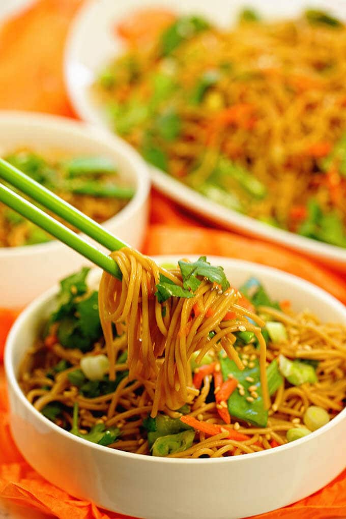 A white bowl filled with teriyaki noodles. There's a pair of green chopsticks that have scooped up a big bite of noodles