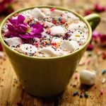 A green cup on a wooden board filled with crock pot hot chocolate, topped with sprinkles!