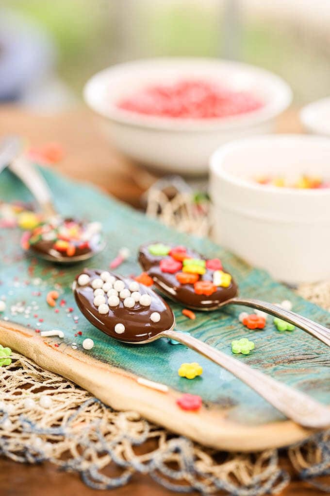 Four Chocolate dipped spoons on blue platter surrounded and topped with sprinkles.