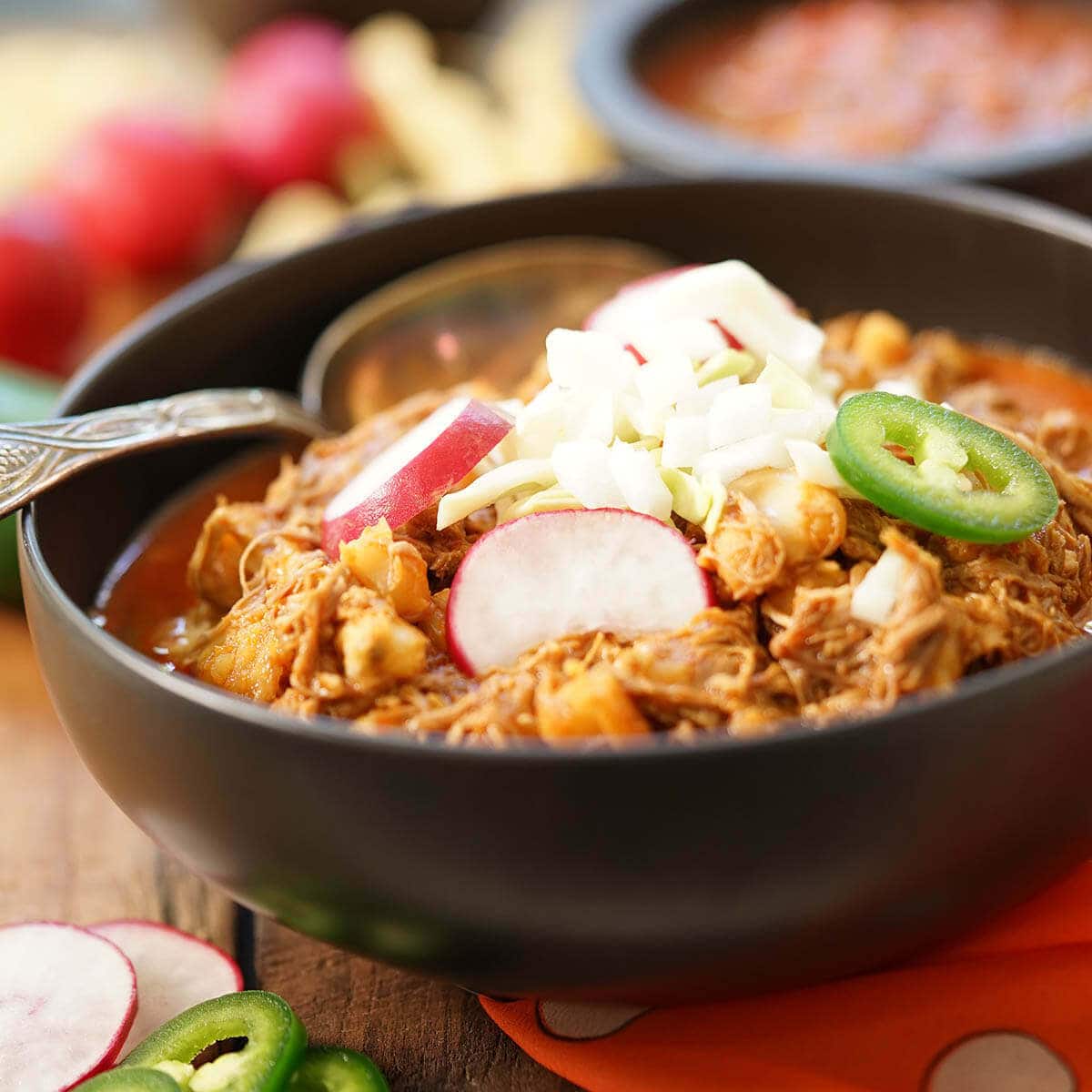 Authentic Posole Recipe in black bowl topped with onions, jalapeños and sliced radishes.