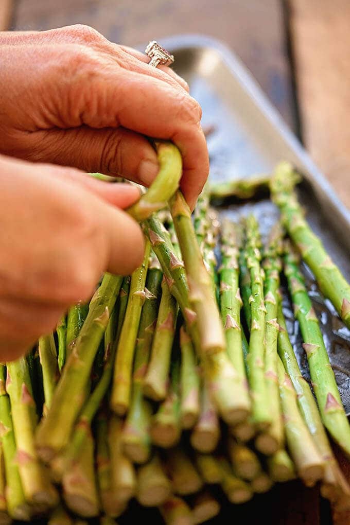 A brunch of asparagus on a sheet pan. The end of one stalk is being snapped off.