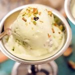 Pistachio Ice Cream in silver cup with spoon. Ice cream is topped with chopped pistachios.