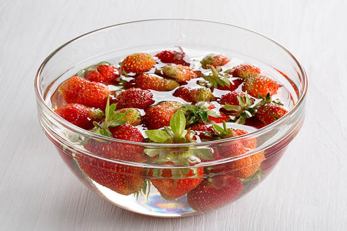 First step in how to make jam is to clean the fruit. Pictured is a clear glass jar filled with strawberries and water, vinegar cleaning solution.