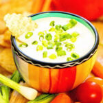 Onion dip in aa colorful bowl topped with sliced green onions and surrounded by chips and vegetables for dipping.