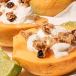 Golden papaya breakfast bowl topped with thick greek yogurt, crunchy granola surround by wedges of lime.