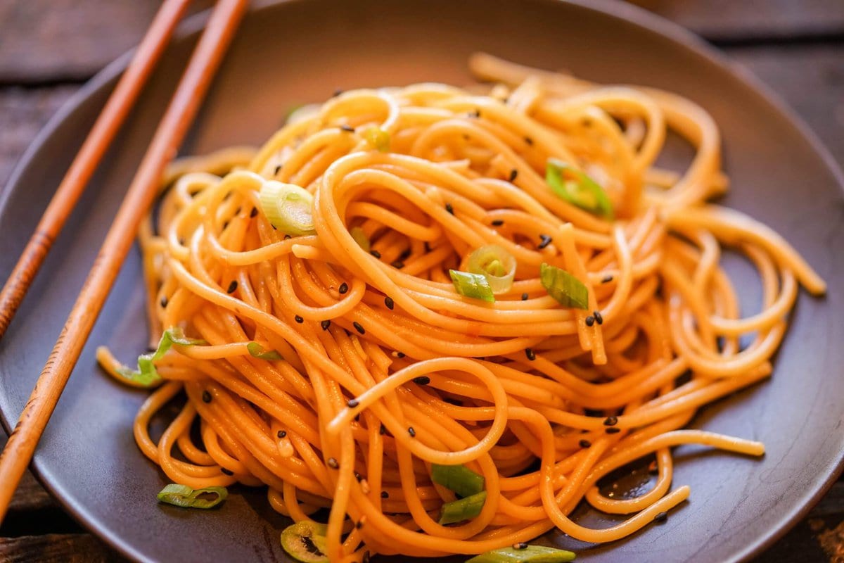 Asian noodles topped with a fiery sauce on a black plate served with chopsticks.