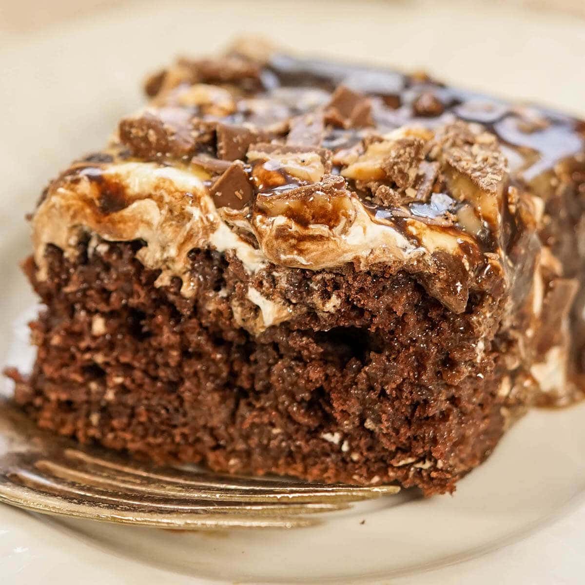 Chocolate cake with whipped cream frosting, topped with crunched up chocolate bars on plate with fork.