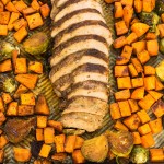 Sliced pork tenderloin on a sheet pan surrounded by diced sweet potatoes and brussels sprouts.