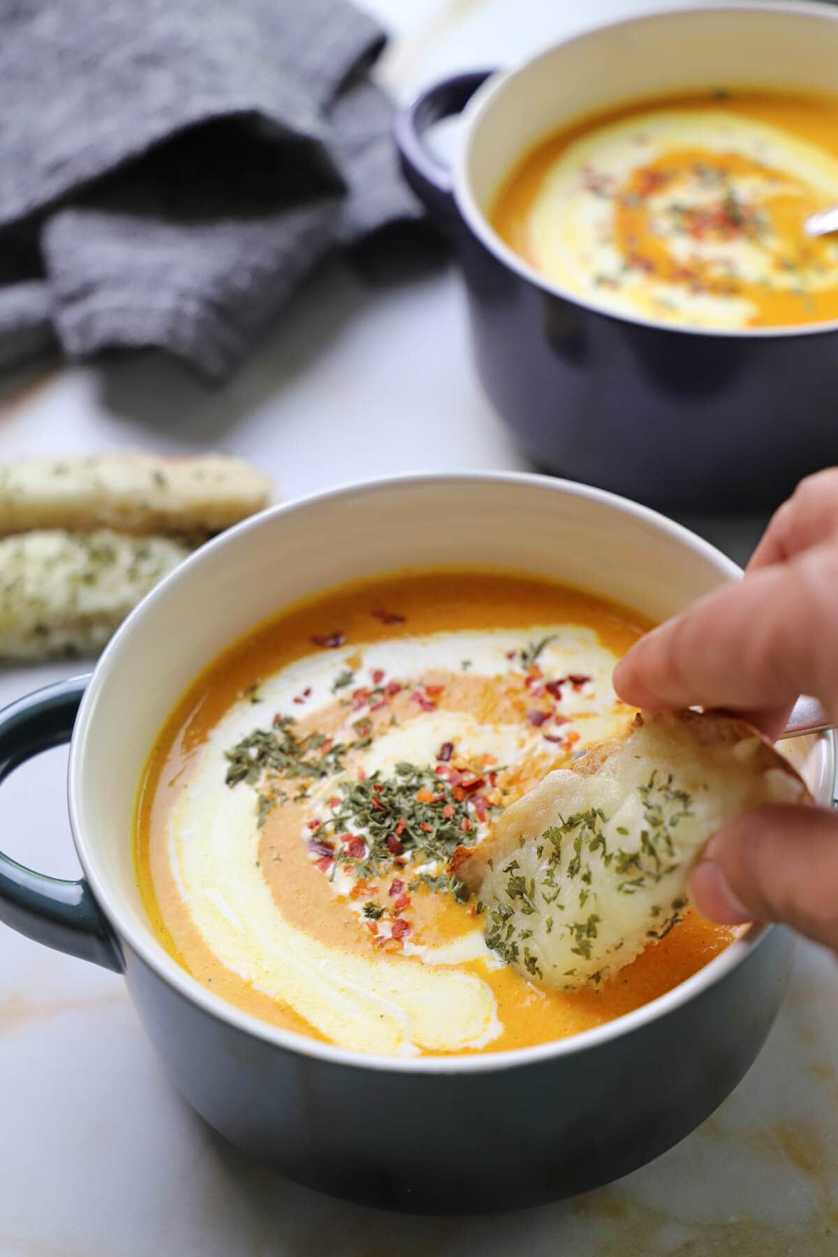 A hand dipping a piece of garlic bread into a bowl of carrot soup.