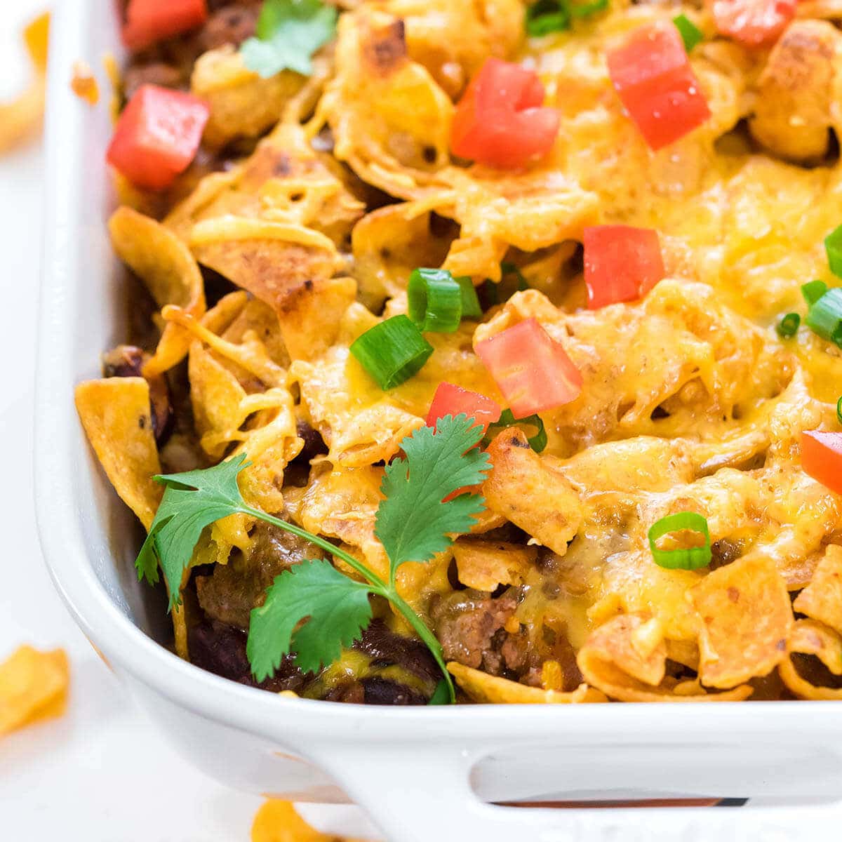 Frito pie in white casserole dish, topped with cheese, tomatoes, green onions and cilantro.