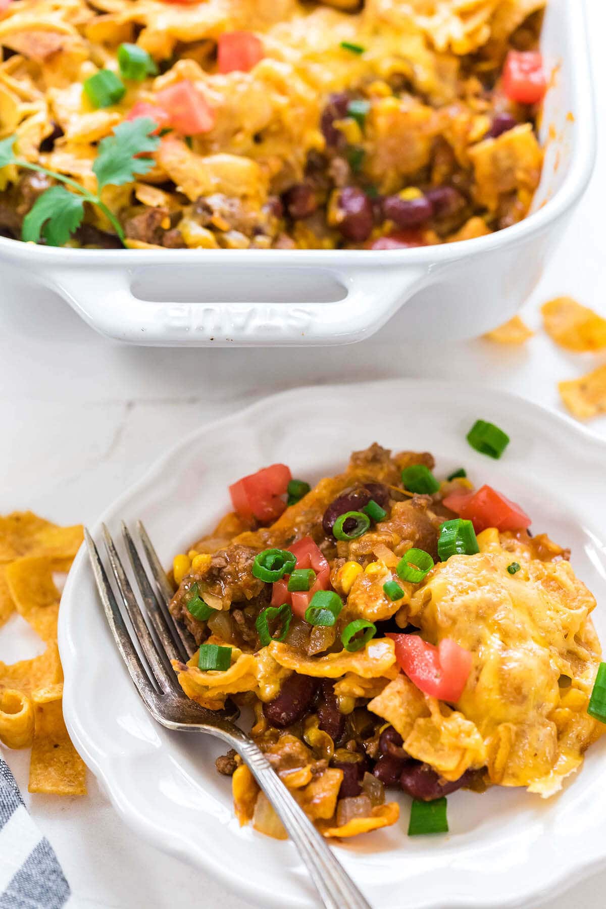 Beef casserole on white plate surrounded by corn chips with a fork to the side. The casserole is pictured in the photo as well.