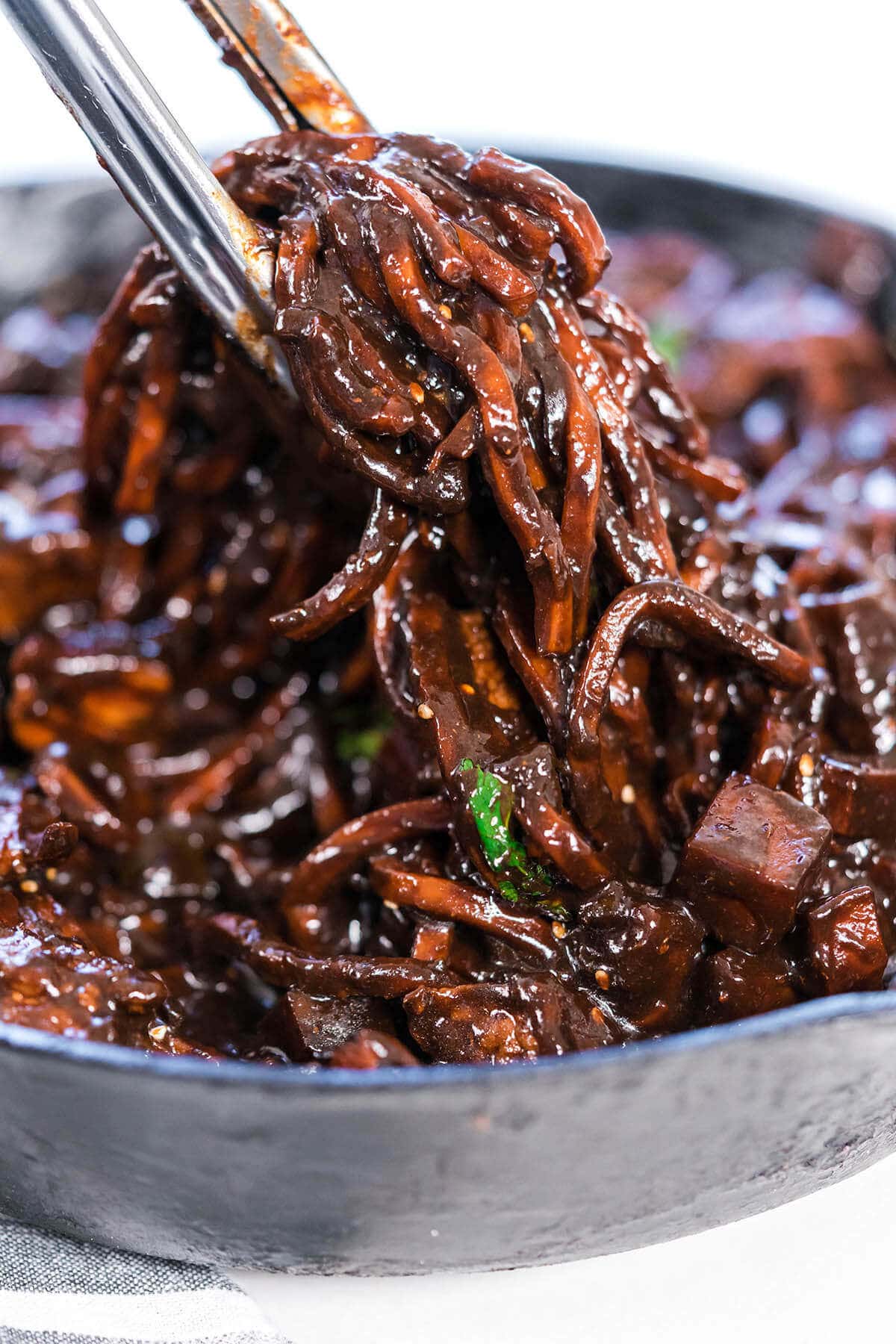 Jjajangmyeon noodles in a skillet being stirred with a pair of tongs.