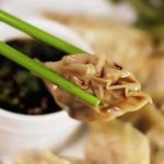 Potsticker held by chopsticks with soy sauce dip in the background.