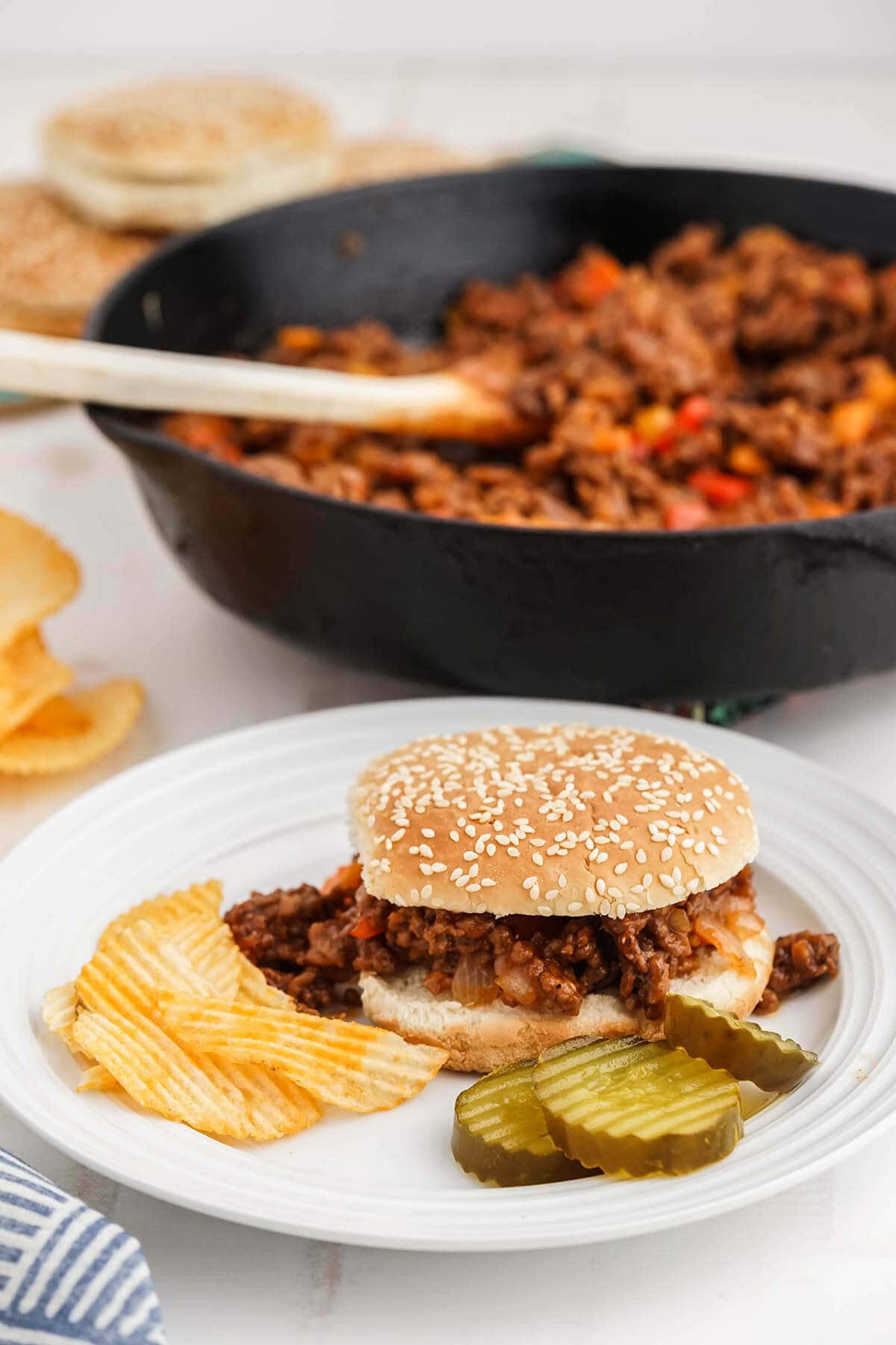 Bun filled with old fashioned sloppy Joes recipe on plate with pickles and potato chips. With skillet in background.