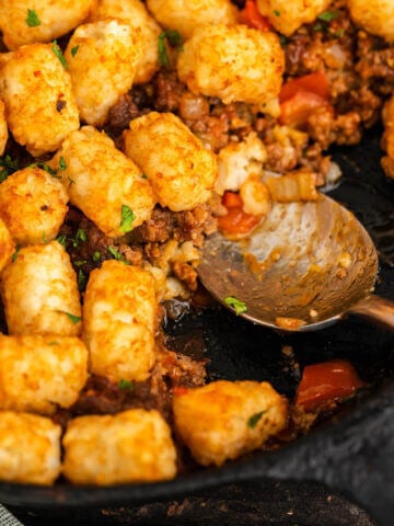Casserole in skillet topped with tater tots. With serving spoon.