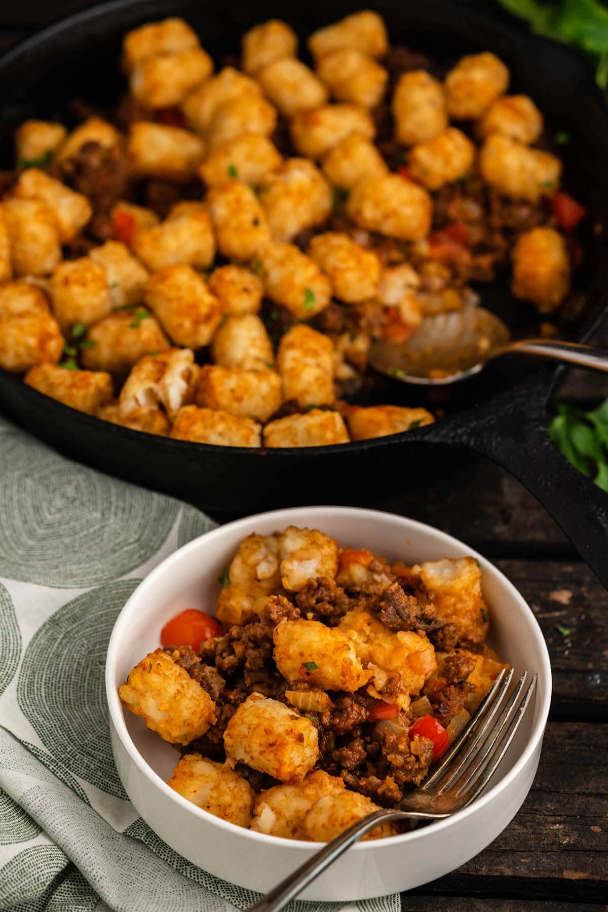 Dish filled with sloppy Joe tater tot casserole in bowl with serving spoon. With skillet filled with meal behind it.