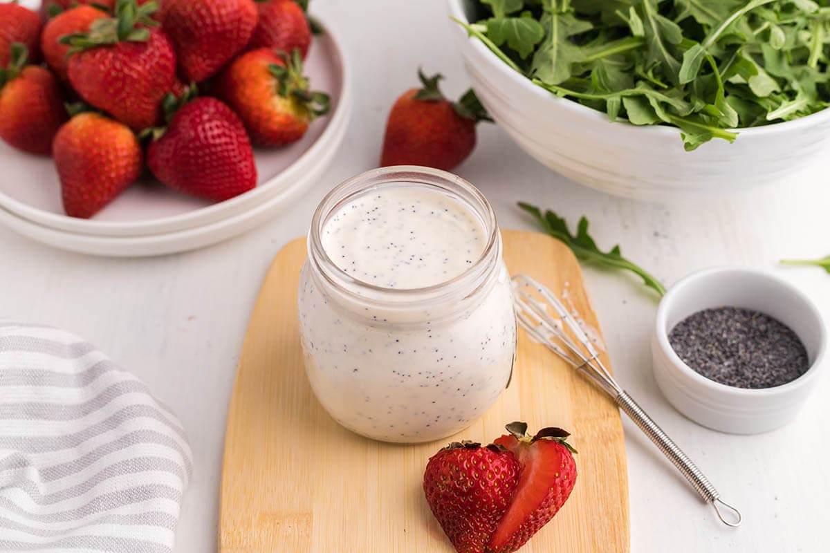 Salad dressing on table with bowl of strawberries and green salad. 